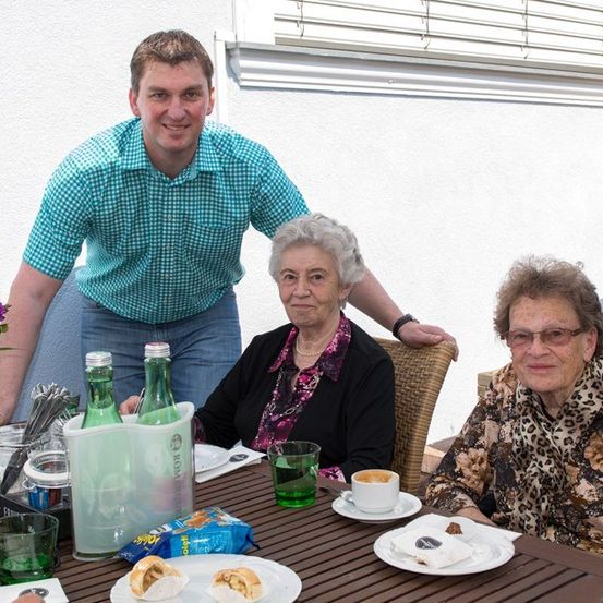 Ein Mann und zwei ältere Frauen sitzen lächelnd an einem Tisch. Der Tisch ist mit Tellern, Tassen und Snacks gedeckt. Hinter ihnen ist eine weiße Wand und ein Fenster zu sehen.