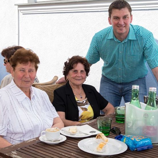Zwei ältere Frauen und ein Mann sitzen lächelnd an einem Tisch. Die Frau links trägt ein weißes Shirt, die Frau in der Mitte eine schwarze Jacke und der Mann ein blau kariertes Hemd. Der Tisch ist mit Tellern, Tassen und Flaschen gedeckt.
