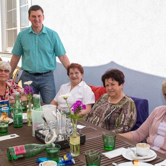 Eine Gruppe von Frauen und einem Mann sitzen um einen Tisch herum und lächeln. Der Tisch ist mit Tellern, Tassen und Blumen gedeckt. Ein Mann steht hinter den Frauen.