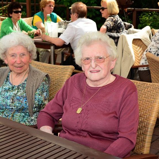 Zwei ältere Frauen sitzen an einem Tisch in einem Restaurant im Freien, lächeln und posieren für ein Foto. Hinter ihnen sitzen andere Leute an Tischen, einige unterhalten sich.