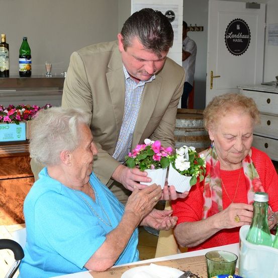 Ein Mann in einem Anzug überreicht zwei älteren Frauen Blumen, die an einem Tisch in einem Essbereich sitzen.