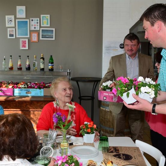 Ein Mann überreicht einer älteren Frau, die mit anderen an einem Tisch sitzt, Blumen in einem Essbereich.