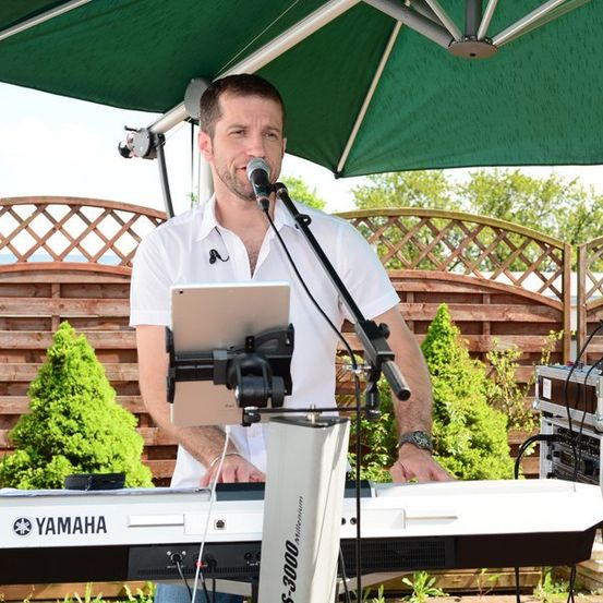 Ein Mann spielt ein Yamaha-Keyboard unter einem grünen Regenschirm, mit einem Mikrofon und einem Laptop auf einem Ständer vor ihm.