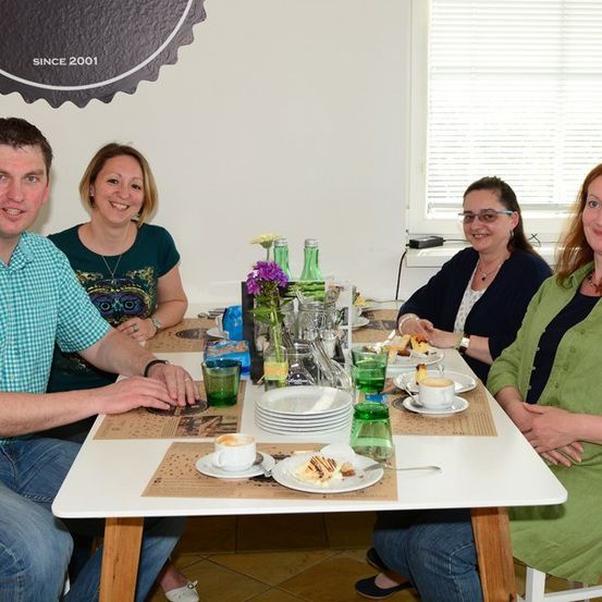 Vier Personen sitzen um einen Tisch mit Tellern, Tassen und einer Blumenvase. Der Mann links lächelt und schaut in die Kamera. Die Frau rechts trägt ein grünes Hemd und hat die Hände auf dem Bauch. Die Frau in der Mitte trägt eine Brille und schaut in die Kamera. Die Frau links trägt eine Halskette und ein Armband.
