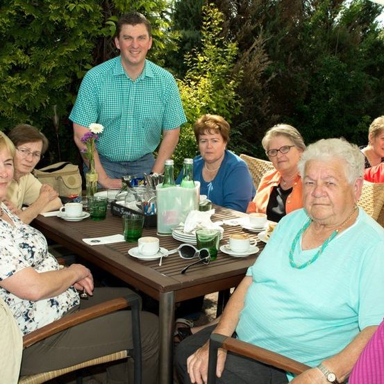 Eine Gruppe älterer Frauen und ein Mann sitzen um einen Tisch in einer Außenumgebung, lächeln und posieren für ein Foto. Der Tisch ist mit Tassen, Tellern und einer Blumenvase gedeckt. Der Mann steht hinter dem Tisch, und die Frauen tragen Brillen und Halsketten.