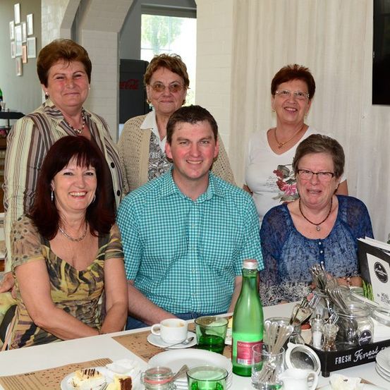 Eine Gruppe lächelnder Erwachsener, darunter zwei Frauen mit Brille, sitzen um einen Tisch mit Essen und Getränken, möglicherweise in einem Restaurant.