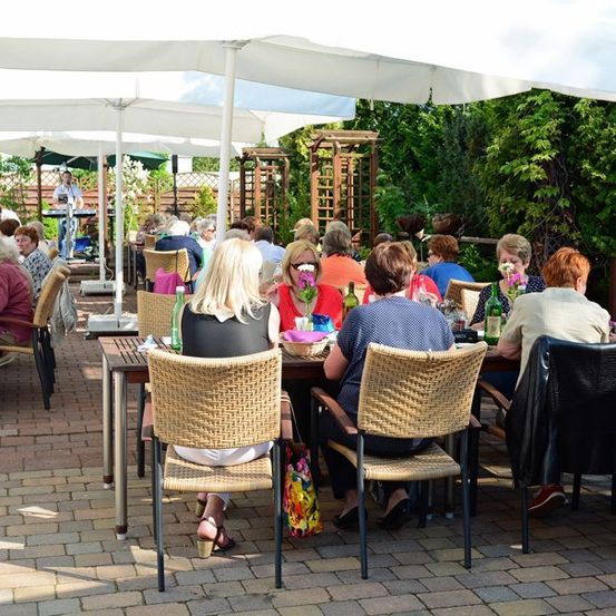 Eine Gruppe von Menschen sitzt an Tischen in einem Restaurant im Freien mit Sonnenschirmen und genießt ihr Essen. Eine Person steht in der Nähe eines Tisches mit einem Mikrofon.