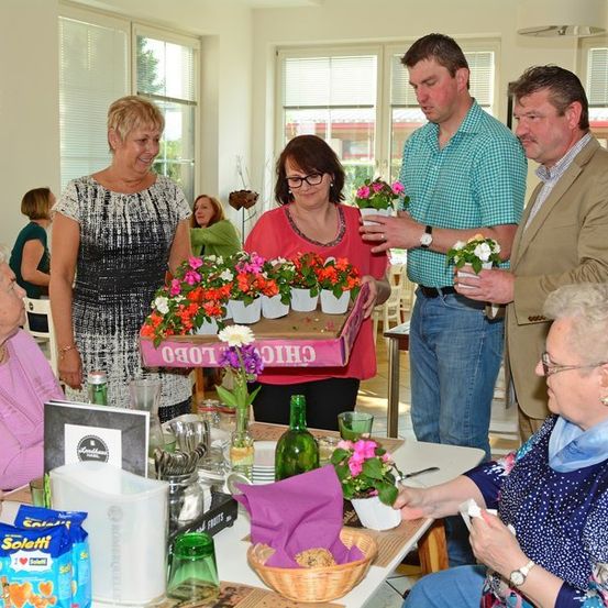 Eine Gruppe von Menschen, darunter ältere Frauen, versammelt sich um einen Tisch mit Blumen, möglicherweise bei einer Veranstaltung in einem Altersheim. Zwei Männer präsentieren den Frauen Blumenarrangements.