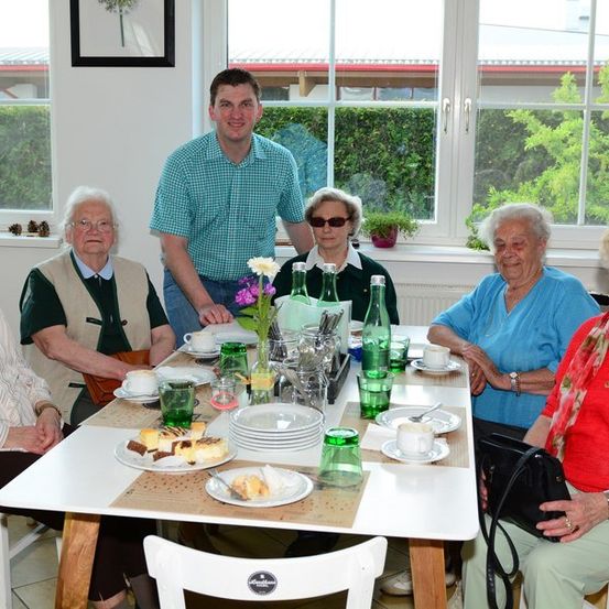 Eine Gruppe älterer Frauen und ein Mann sitzen um einen Tisch mit Tellern, Tassen und Blumen. Sie lächeln und scheinen ihre Mahlzeit zu genießen. Der Tisch ist mit grünen Gläsern und einer Vase mit Blumen gedeckt. Der Raum hat große Fenster mit Pflanzen draußen.