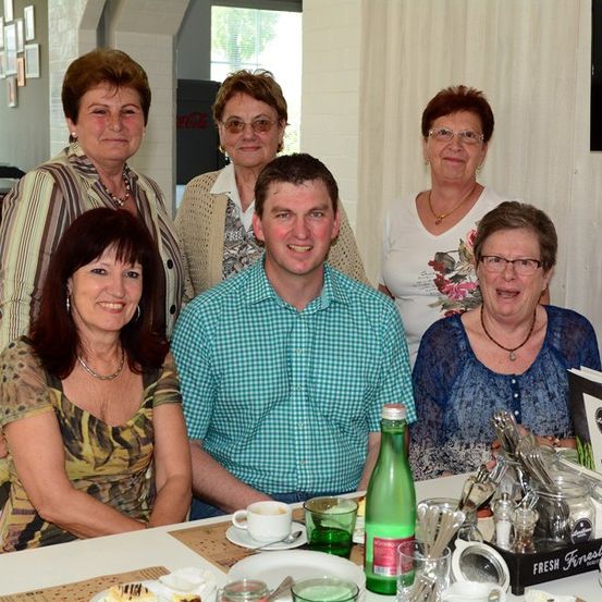 Eine Gruppe von Menschen sitzt in einem Restaurant um einen Tisch herum, lächelt und posiert für ein Foto. Auf dem Tisch stehen Teller, Tassen und Flaschen. Dahinter steht ein Fass, eine Pflanze und ein Fernseher.