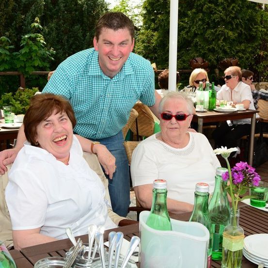 Zwei ältere Frauen und ein Mann sitzen an einem Tisch in einem Restaurant im Freien. Der Mann steht hinter der Frau auf der linken Seite, und sie alle lächeln. Auf dem Tisch stehen Flaschen, Teller und Blumen.