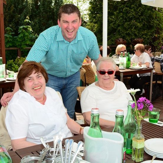 Eine Gruppe von Menschen sitzt an einem Tisch in einem Restaurant im Freien, wobei ein Mann hinter zwei lächelnden Frauen steht. Der Tisch ist mit Tellern, Gläsern und Blumen gedeckt. Im Hintergrund sitzen andere Menschen an Tischen, und das Gebiet ist von Bäumen und Pflanzen umgeben.