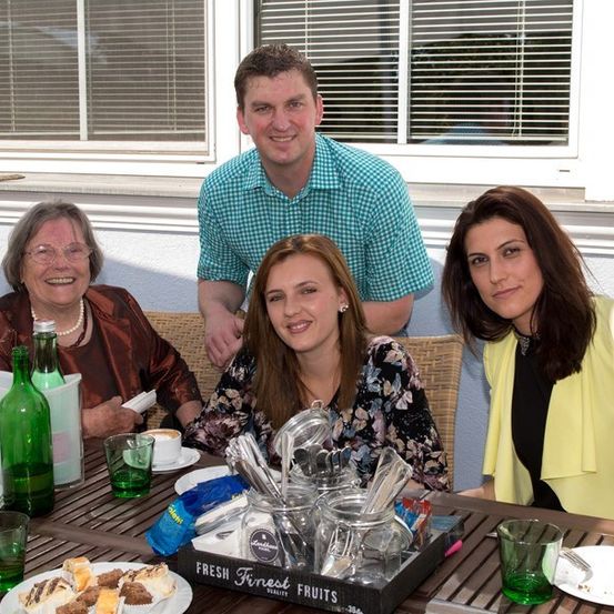 Vier Menschen sitzen bei einem Foto mit Essen und Getränken am Tisch und lächeln. Eine Frau links trägt eine Brille und eine Halskette. Ein Mann in einem blauen Hemd steht hinter einer Frau in einem Blumenhemd. Eine Frau in einem gelben Blazer sitzt rechts.