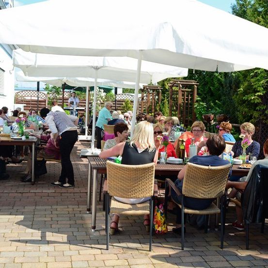 Eine Gruppe von Menschen isst im Freien unter weißen Sonnenschirmen auf einer Ziegelterrasse. Die Tische sind mit Tellern, Gläsern und Blumen gedeckt. Eine Frau in einem schwarzen Kleid steht in der Nähe der Tische.