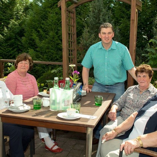 Eine Gruppe älterer Frauen und ein Mann sitzen um einen Tisch in einem Garten. Der Tisch hat Teller, Tassen und Flaschen. Der Mann steht hinter dem Tisch.