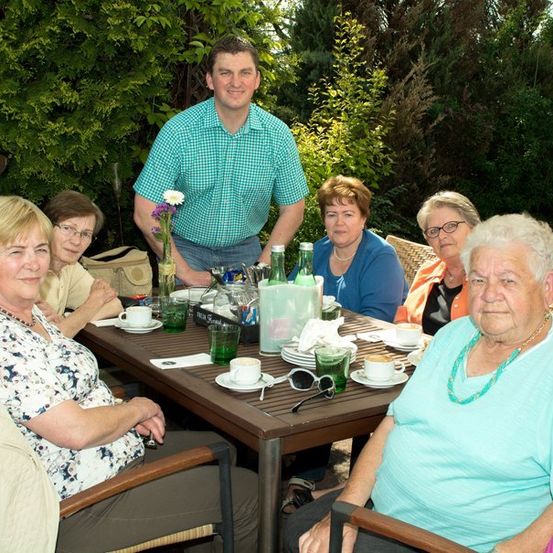 Eine Gruppe älterer Frauen und ein Mann sitzen um einen Tisch in einer Außenumgebung. Der Tisch ist mit Tassen, Tellern und einem Blumenvase gedeckt. Der Mann steht hinter dem Tisch und lächelt.