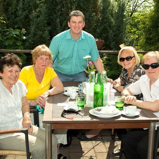 Fünf Personen, darunter ein Mann in einem grünen Hemd, sitzen um einen Tisch in einem Garten im Freien, lächeln und posieren für ein Foto. Der Tisch ist mit Tellern, Tassen und Flaschen gedeckt.