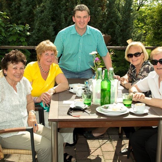 Fünf Personen sitzen um einen Tisch in einer Außenumgebung, lächeln und posieren für ein Foto. Der Tisch ist mit Tellern, Tassen und Flaschen gedeckt. Im Hintergrund befinden sich Grünflächen und ein Zaun.