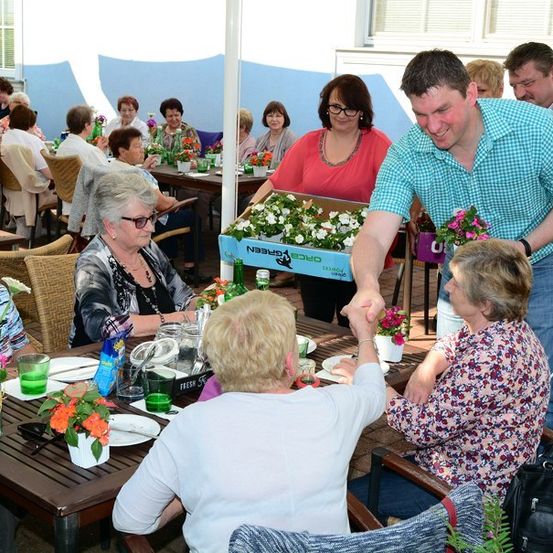 Eine Gruppe älterer Frauen sitzt in einem Restaurant um einen Tisch herum, wobei ein Mann einer von ihnen Blumen präsentiert.