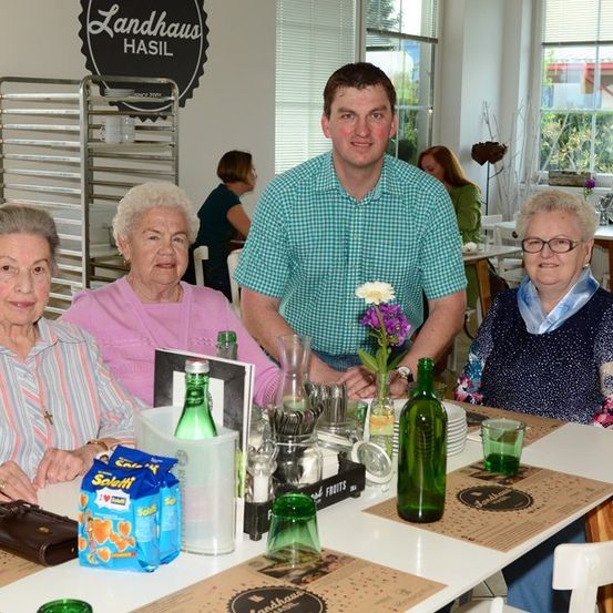 Drei ältere Frauen und ein Mann sitzen an einem Tisch in einem Restaurant mit einem Schild, das 'Landhaus Hasil' steht. Sie sind umgeben von Flaschen, einer Blume und einer Tüte Snacks.