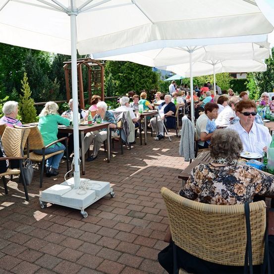 Eine Gruppe älterer Frauen essen im Freien unter großen weißen Sonnenschirmen auf einer Ziegelterrasse.