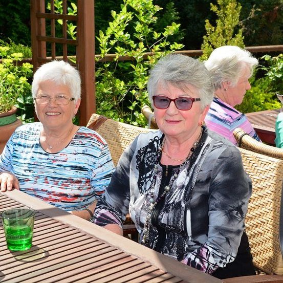 Zwei ältere Frauen sitzen an einem Tisch in einem Restaurant im Freien, lächeln und schauen in die Kamera. Eine trägt eine Brille und eine Halskette, die andere eine Sonnenbrille. Ein Topf mit Pflanzen und ein grünes Getränk stehen auf dem Tisch.