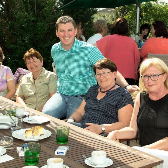 Eine Gruppe von Menschen, darunter ein Mann und vier Frauen, sitzt um einen Tisch in einer Außenumgebung und posiert für ein Foto. Der Tisch ist mit Tellern, Tassen und einer Flasche gedeckt. Hinter ihnen sitzen und stehen weitere Personen, und ein grüner Schirm ist sichtbar.