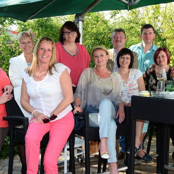 Eine Gruppe von Menschen posiert für ein Foto unter einem grünen Schirm, einige sitzen und andere stehen. Sie sind lässig gekleidet und lächeln, mit einem Tisch voller Weingläser und Teller im Vordergrund.