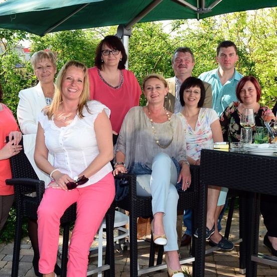 Eine Gruppe von Menschen posiert für ein Foto in einer Außenumgebung, einige sitzen auf Stühlen unter einem Schirm, mit einem Tisch, der Weingläser und Teller im Vordergrund hält.