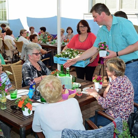 Eine Gruppe älterer Frauen sitzt um einen Tisch in einem Garten, ein Mann verteilt Blumen.