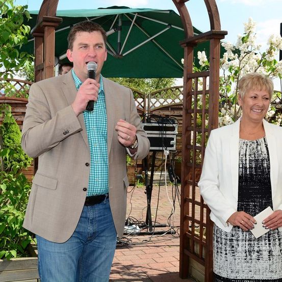 Ein Mann spricht in ein Mikrofon, während eine Frau neben ihm steht und beide lächeln, in einer Gartenumgebung.