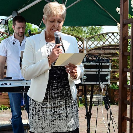 Eine Frau steht unter einem grünen Schirm, hält ein Papier und ein Mikrofon in der Hand, während ein Mann im Hintergrund Klavier spielt.