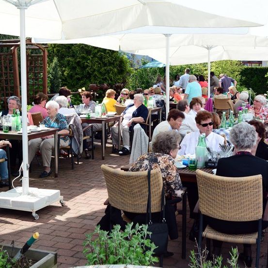 Eine Gruppe älterer Menschen sitzt an Tischen in einem Außenrestaurant, einige unter Schirmen, mit umliegenden Grünpflanzen.