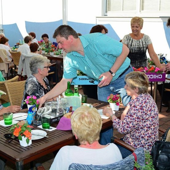 Eine Gruppe älterer Frauen und ein Mann sitzen um einen Tisch in einem Restaurant im Freien, ein Mann steht und serviert ihnen.