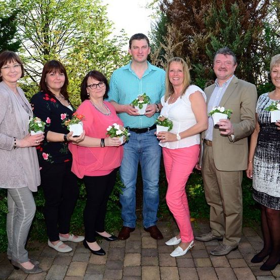Eine Gruppe von Erwachsenen, sowohl Männer als auch Frauen, posiert für ein Foto in einem Garten mit Topfpflanzen in den Händen.