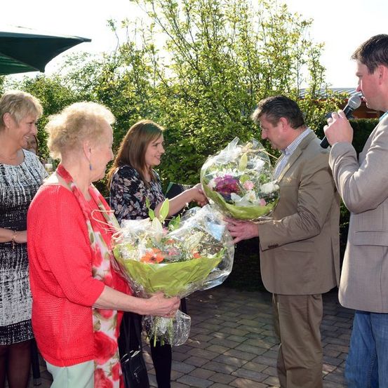 Eine Gruppe von Menschen steht im Freien, eine Frau in einem roten Pullover hält einen Blumenstrauß, eine andere Frau hält einen Strauß und ein Mann mit Mikrofon spricht zu ihnen.