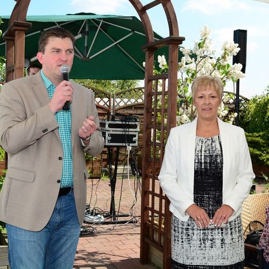 Ein Mann und eine Frau stehen unter einem Holzbogen in einem Garten, wobei der Mann in ein Mikrofon spricht. Die Frau steht neben ihm, und ein Lautsprecher ist im Hintergrund sichtbar.