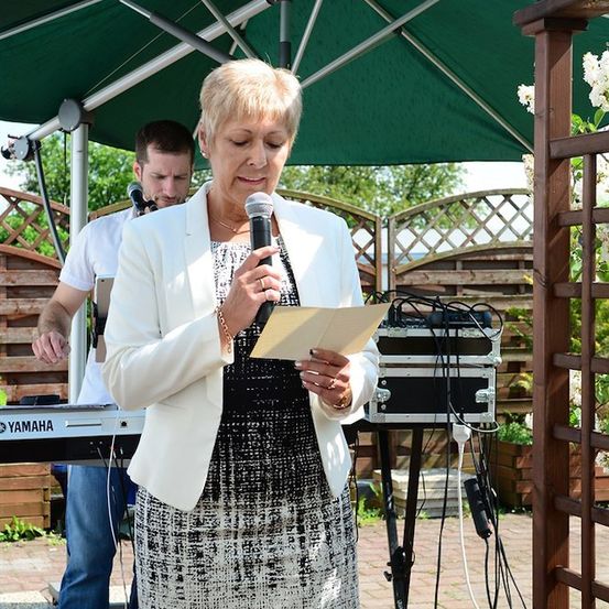 Eine Frau in einer weißen Jacke und einem schwarzen Kleid steht unter einem grünen Schirm, hält ein Mikrofon und liest von einem Stück Papier. Ein Mann spielt hinter ihr das Keyboard.