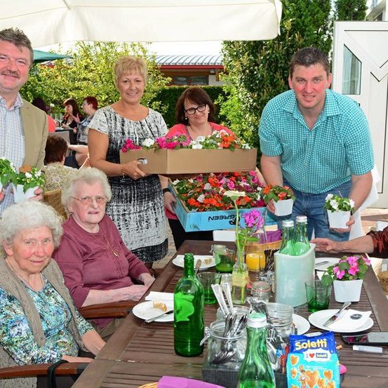 Eine Gruppe von Menschen, einschließlich älterer Frauen, versammelt sich um einen Tisch in einer Außenumgebung. Sie halten Blumentöpfe und lächeln. Der Tisch ist mit Tellern, Gläsern und verschiedenen Gegenständen gedeckt. Im Hintergrund ist ein Garten mit Bäumen und einem Gebäude zu sehen.