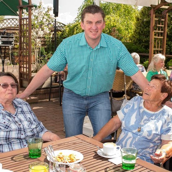 Ein Mann steht hinter zwei älteren Frauen, die an einem Tisch in einem Garten sitzen, lächelt und posiert für ein Foto. Auf dem Tisch stehen ein Teller mit Essen, Tassen und eine Untertasse.