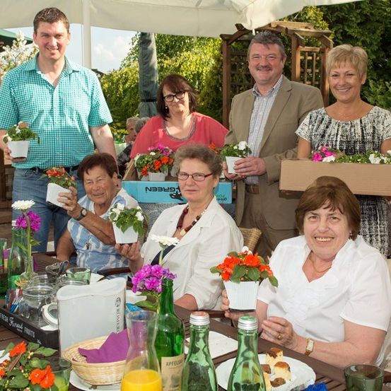 Eine Gruppe von Menschen, sowohl Männer als auch Frauen, versammelt sich um einen Tisch, lächelt und hält Topfpflanzen. Der Tisch ist mit Blumen, Flaschen und einem Krug gedeckt. Sie befinden sich in einem Garten mit Bäumen und einer Holzstruktur im Hintergrund.