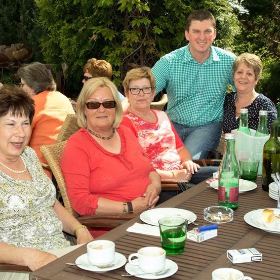 Eine Gruppe von Menschen sitzt an einem Tisch in einem Garten im Freien, lächelt und posiert für ein Foto. Auf dem Tisch stehen Teller, Tassen und Flaschen.