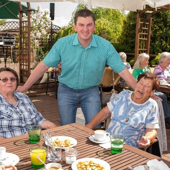 Ein Mann steht hinter zwei älteren Frauen, die an einem Tisch in einem Café im Freien sitzen. Die Frauen lachen, und der Mann lächelt. Es gibt Teller mit Essen, Tassen und Gläser auf dem Tisch. Andere Leute sitzen im Hintergrund an Tischen.