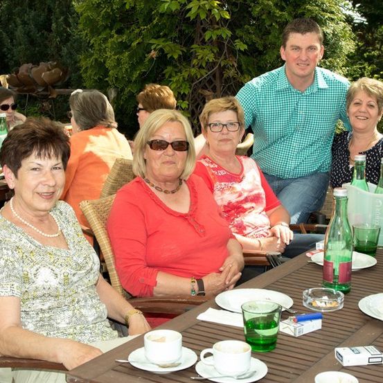 Eine Gruppe von Menschen sitzt um einen Tisch in einem Restaurant im Freien. Sie lächeln und scheinen ihre Mahlzeit zu genießen. Es gibt Teller, Tassen und Flaschen auf dem Tisch. Hinter ihnen sind Bäume und eine Metallskulptur.