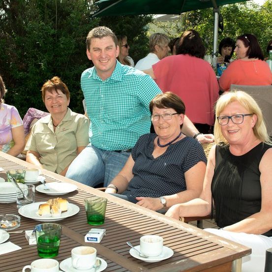 Eine Gruppe Erwachsener, darunter ein Mann in einem blauen Hemd, sitzen um einen Tisch in einer Außenumgebung und lächeln für ein Foto. Der Tisch ist mit Tellern, Tassen und einer Flasche gedeckt. Im Hintergrund sitzen weitere Personen an Tischen unter einem Schirm.