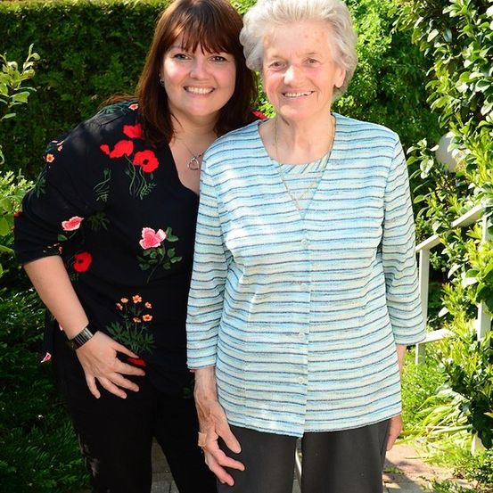 Zwei Frauen stehen in einem Garten, eine mit einem Blumenhemd und die andere mit einem gestreiften Hemd, beide lächeln.