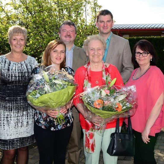 Eine Gruppe von Menschen steht draußen zusammen, zwei Frauen halten Blumensträuße, eine ältere Frau lächelt und zwei Männer stehen hinter ihnen mit einem Gebäude im Hintergrund.