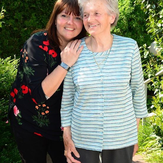 Zwei Frauen, eine ältere und eine jüngere, stehen eng beieinander und lächeln für ein Foto in einem Garten mit grünen Büschen und einem weißen Zaun.