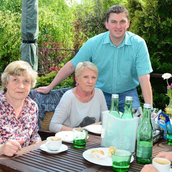 Drei Menschen sitzen an einem Tisch in einem Garten, ein Mann steht hinter ihnen. Sie lächeln und scheinen ihre Mahlzeit zu genießen. Der Tisch ist mit Tellern, Tassen und Flaschen gedeckt. Der Garten ist von grünen Pflanzen und Bäumen umgeben.