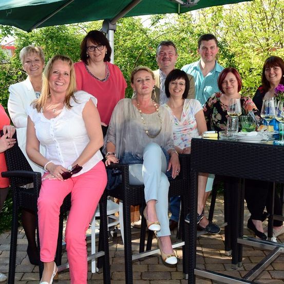 Eine Gruppe von Menschen, darunter Männer und Frauen, posiert für ein Foto in einer Außenumgebung mit einem grünen Schirm und einem Tisch mit Weingläsern.
